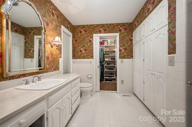 bathroom featuring wallpapered walls, toilet, vanity, wainscoting, and tile walls