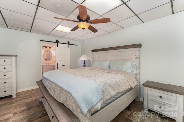 bedroom with a barn door, dark hardwood / wood-style floors, a drop ceiling, ceiling fan, and ensuite bathroom