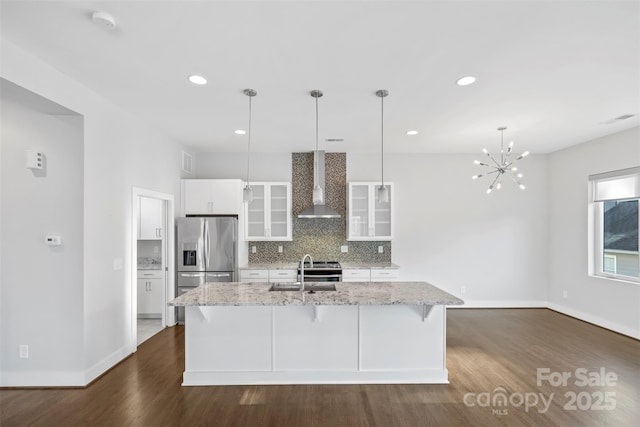 kitchen featuring white cabinets, a center island with sink, wall chimney exhaust hood, stainless steel fridge, and decorative light fixtures