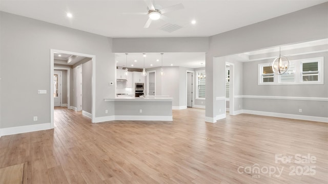 unfurnished living room with light wood-type flooring, visible vents, a raised ceiling, and ceiling fan with notable chandelier