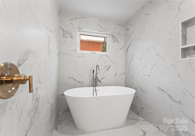 full bath with marble finish floor, a freestanding bath, and stone wall