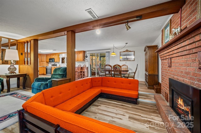 living area with visible vents, light wood-style flooring, a brick fireplace, a textured ceiling, and beamed ceiling