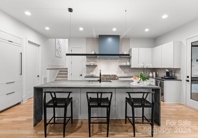 kitchen with sink, light hardwood / wood-style floors, wall chimney exhaust hood, white cabinets, and a center island with sink