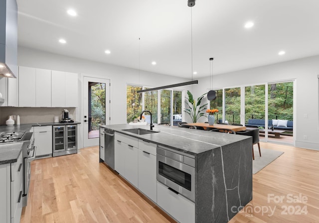 kitchen with an island with sink, hanging light fixtures, sink, beverage cooler, and white cabinets