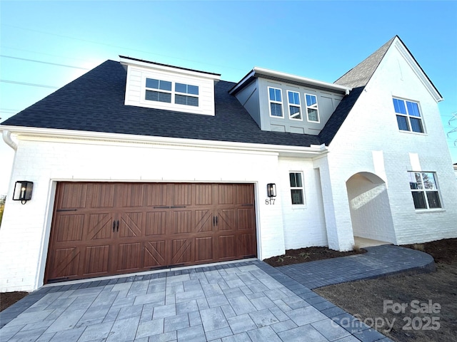 view of front of house with decorative driveway, brick siding, an attached garage, and roof with shingles