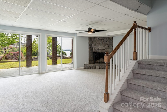 interior space with ceiling fan, a paneled ceiling, carpet flooring, and a stone fireplace