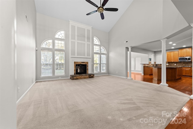 unfurnished living room with ceiling fan, a stone fireplace, decorative columns, a towering ceiling, and light carpet