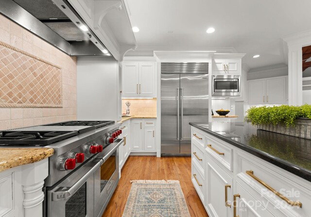 kitchen with white cabinets, exhaust hood, backsplash, dark stone countertops, and built in appliances