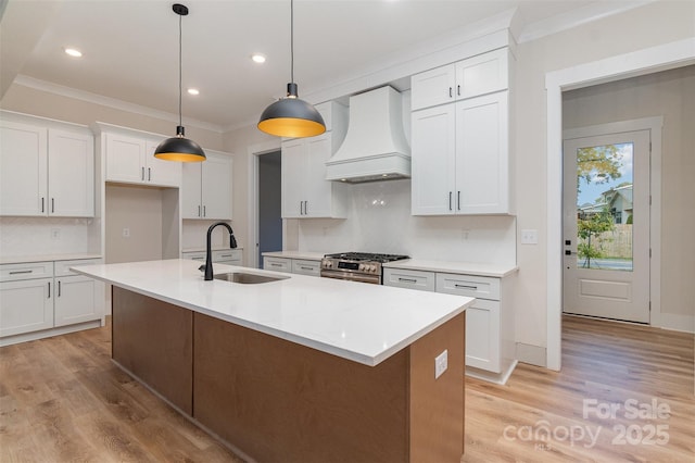 kitchen featuring premium range hood, a center island with sink, sink, hanging light fixtures, and white cabinets