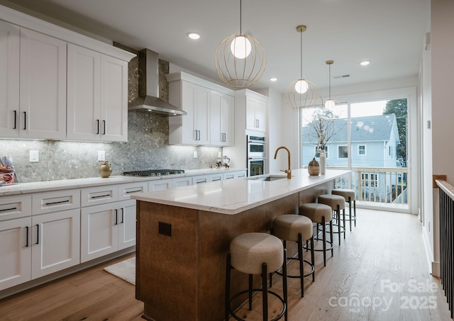 kitchen featuring tasteful backsplash, wall chimney exhaust hood, decorative light fixtures, white cabinetry, and an island with sink