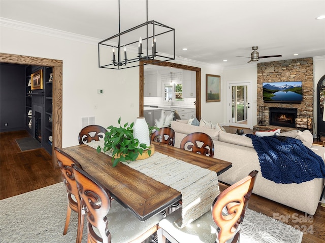 dining room featuring a stone fireplace, crown molding, hardwood / wood-style flooring, and ceiling fan with notable chandelier