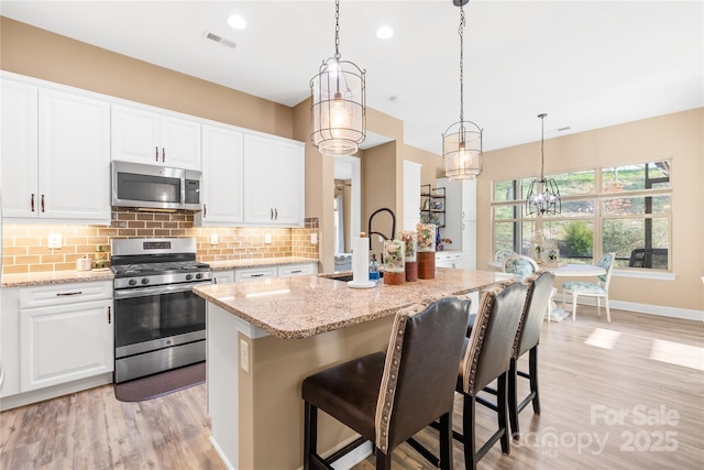 kitchen with appliances with stainless steel finishes, light stone counters, white cabinets, hanging light fixtures, and an island with sink