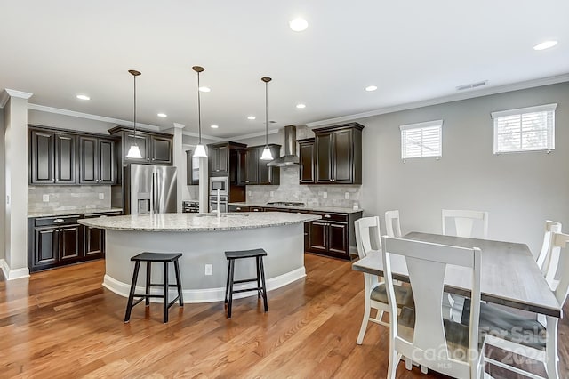 kitchen featuring stainless steel appliances, light wood-type flooring, hanging light fixtures, wall chimney exhaust hood, and a kitchen island with sink