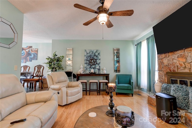 living room featuring light hardwood / wood-style floors, ceiling fan, a textured ceiling, and a stone fireplace