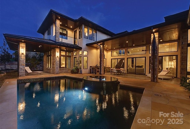 back of house at twilight with metal roof, a balcony, a patio area, stone siding, and a standing seam roof