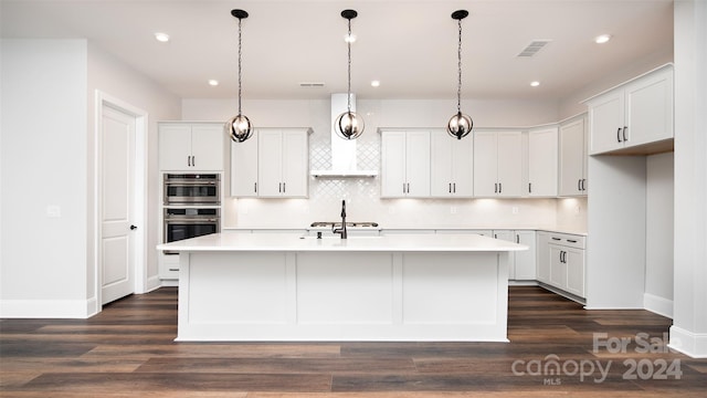 kitchen featuring white cabinetry, wall chimney range hood, dark hardwood / wood-style floors, double oven, and an island with sink