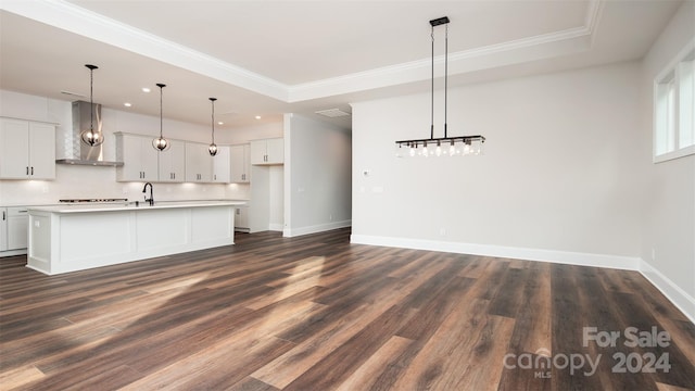 kitchen with dark wood-type flooring, wall chimney range hood, crown molding, an island with sink, and decorative light fixtures