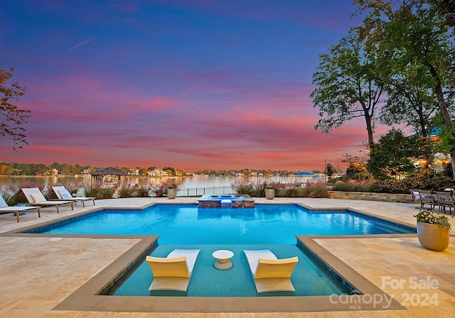 pool at dusk featuring an in ground hot tub, a patio, and a water view