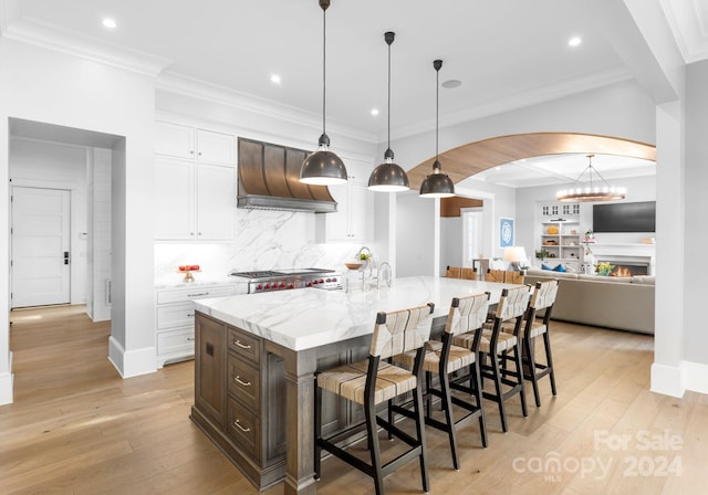 kitchen featuring light wood-type flooring, premium range hood, decorative light fixtures, white cabinets, and an island with sink