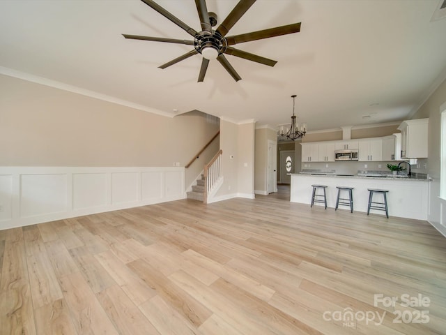 unfurnished living room with light wood-type flooring, sink, ceiling fan with notable chandelier, and ornamental molding