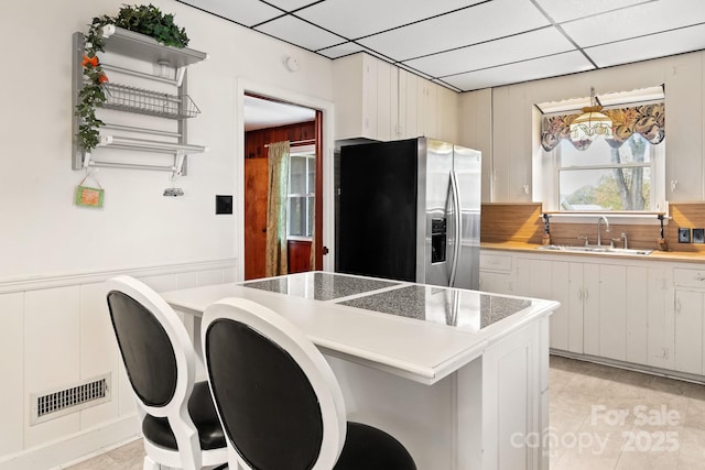 kitchen featuring sink, stainless steel fridge, a paneled ceiling, wooden walls, and white cabinets