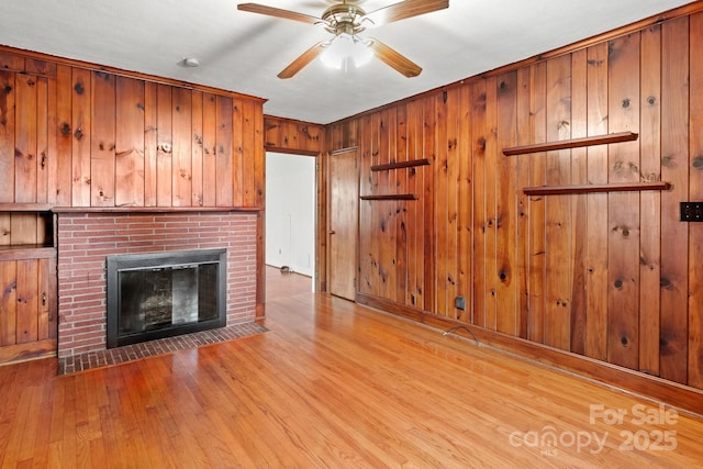 unfurnished living room with wood walls, ceiling fan, light hardwood / wood-style floors, and a brick fireplace