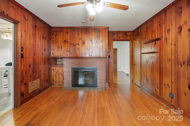 unfurnished living room with a brick fireplace, ceiling fan, wooden walls, crown molding, and light hardwood / wood-style flooring