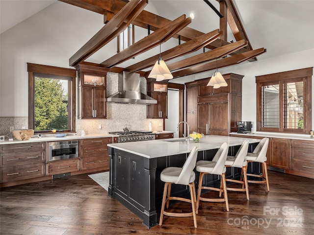 kitchen with sink, wall chimney exhaust hood, dark hardwood / wood-style flooring, backsplash, and a center island with sink