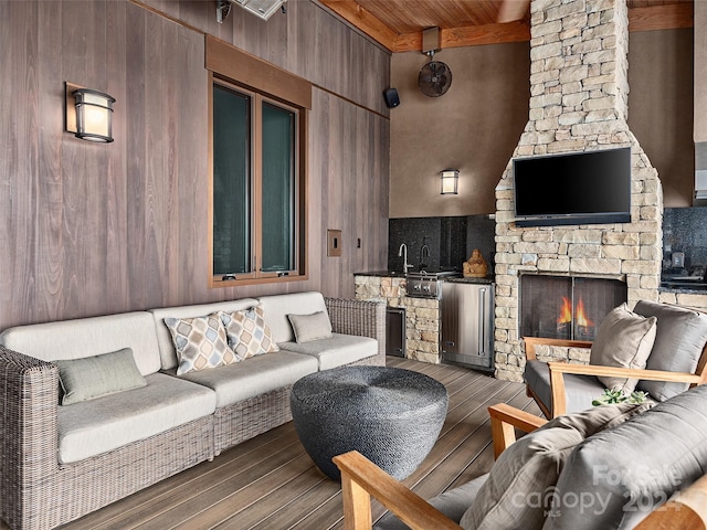 living room with beam ceiling, wooden ceiling, dark wood-type flooring, a stone fireplace, and wood walls