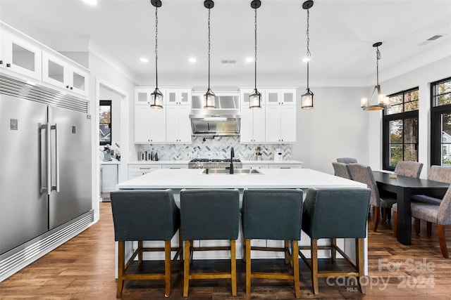 kitchen with sink, wall chimney range hood, dark hardwood / wood-style flooring, built in refrigerator, and an island with sink