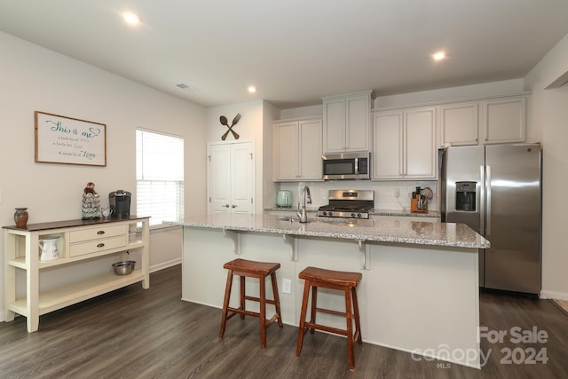 kitchen with stainless steel appliances, an island with sink, dark wood-type flooring, and light stone counters