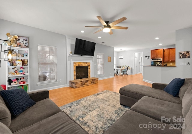 living room featuring ceiling fan with notable chandelier, light wood-type flooring, and a stone fireplace