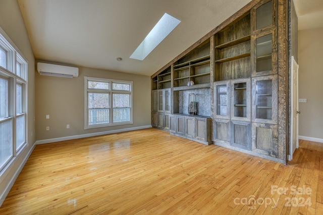 unfurnished room featuring built in shelves, light wood-type flooring, a wall mounted air conditioner, and vaulted ceiling with skylight