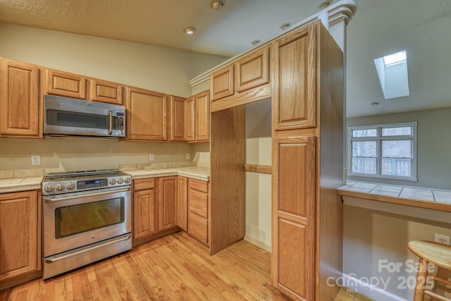 kitchen with tile counters, appliances with stainless steel finishes, lofted ceiling with skylight, and light hardwood / wood-style floors