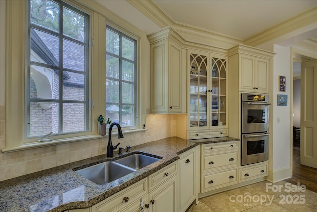 kitchen featuring stainless steel double oven, decorative backsplash, dark stone counters, cream cabinets, and sink