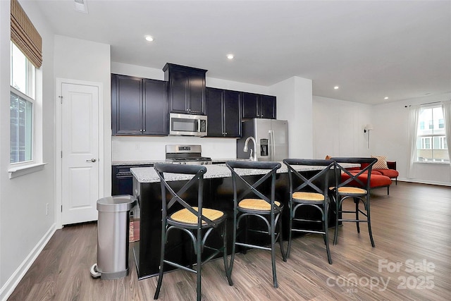 kitchen featuring a breakfast bar, stainless steel appliances, light stone counters, wood-type flooring, and an island with sink