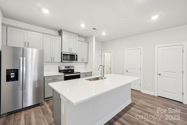 kitchen with sink, wood-type flooring, an island with sink, stainless steel appliances, and decorative backsplash