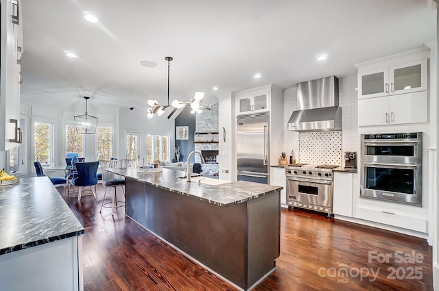 kitchen featuring high end appliances, wall chimney range hood, a sink, a chandelier, and tasteful backsplash