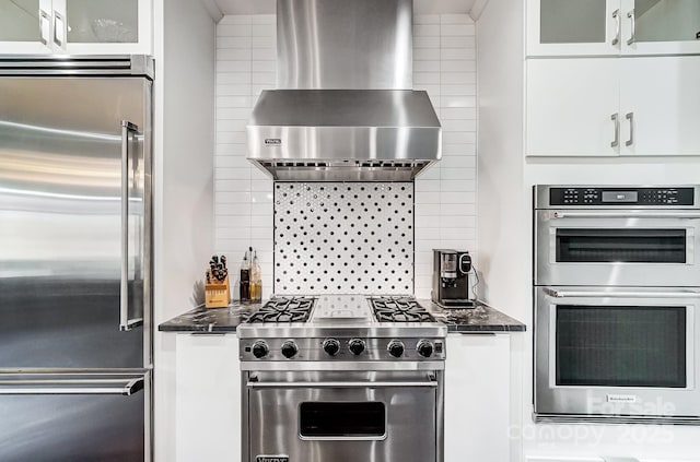 kitchen featuring wall chimney exhaust hood, backsplash, white cabinetry, glass insert cabinets, and high quality appliances