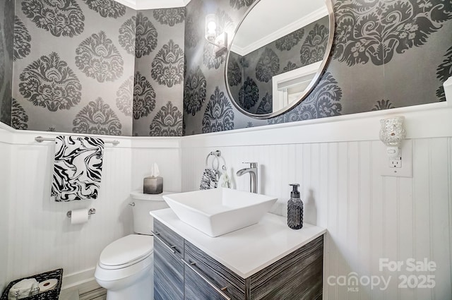 half bath with a decorative wall, wallpapered walls, wainscoting, vanity, and toilet