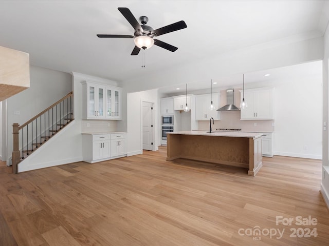 kitchen with white cabinetry, light wood-type flooring, decorative light fixtures, an island with sink, and wall chimney exhaust hood