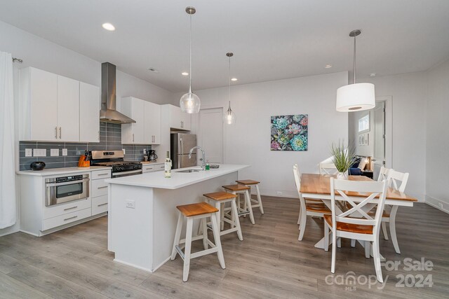 kitchen with stainless steel appliances, white cabinetry, wall chimney range hood, decorative light fixtures, and an island with sink