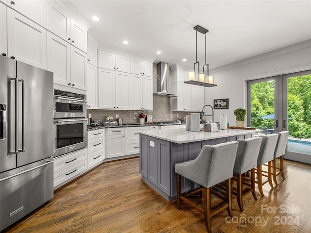 kitchen featuring stainless steel appliances, light stone countertops, wall chimney exhaust hood, a kitchen island with sink, and white cabinets