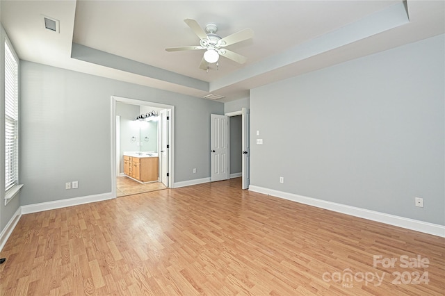 unfurnished bedroom with a raised ceiling, ensuite bathroom, ceiling fan, and light wood-type flooring