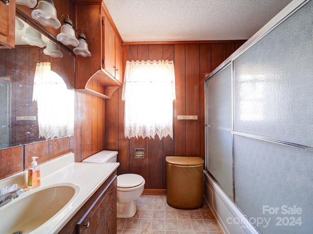 full bathroom featuring vanity, wooden walls, toilet, enclosed tub / shower combo, and a textured ceiling