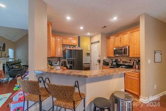 kitchen with kitchen peninsula, appliances with stainless steel finishes, light brown cabinetry, vaulted ceiling, and a breakfast bar area