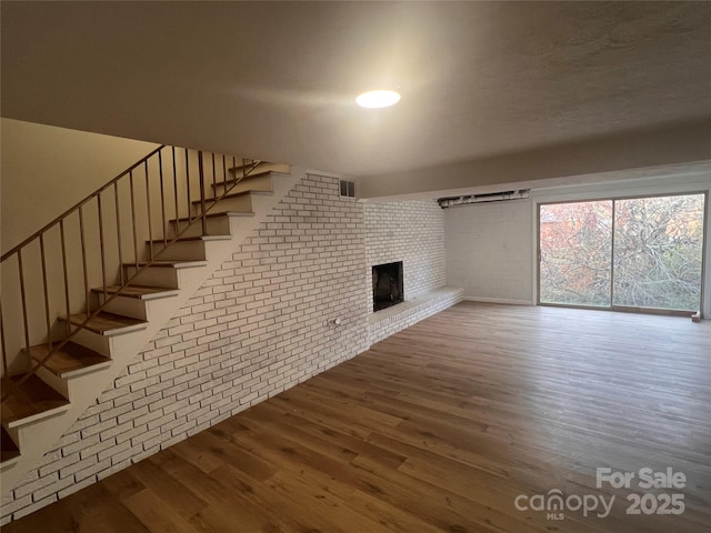 unfurnished living room featuring brick wall, visible vents, a fireplace, wood finished floors, and stairway