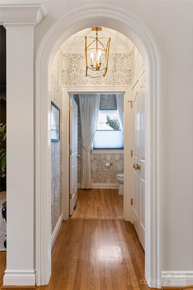 hall featuring arched walkways, wood-type flooring, an inviting chandelier, ornamental molding, and wallpapered walls