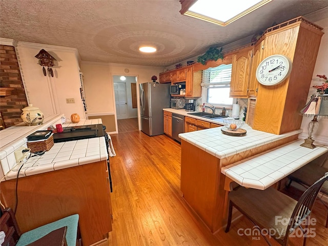 kitchen with tile counters, brown cabinetry, appliances with stainless steel finishes, a peninsula, and a sink