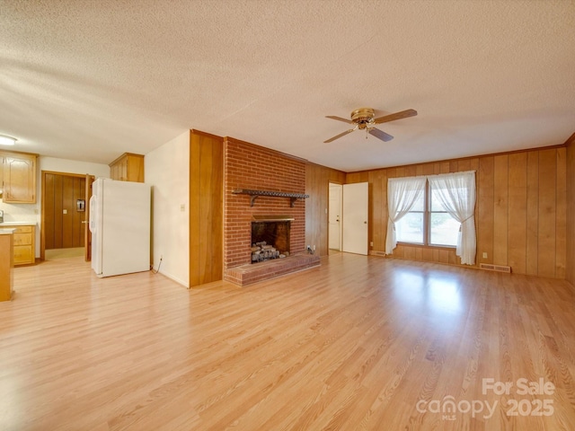 unfurnished living room featuring wood walls, a brick fireplace, ceiling fan, a textured ceiling, and light hardwood / wood-style floors